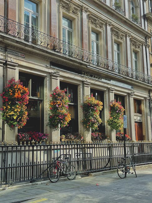 A floral display outside a historic building with ornate stone window frames and decorative iron balconies, featuring large hanging baskets filled with vibrant pink, orange, and yellow flowers. The bo