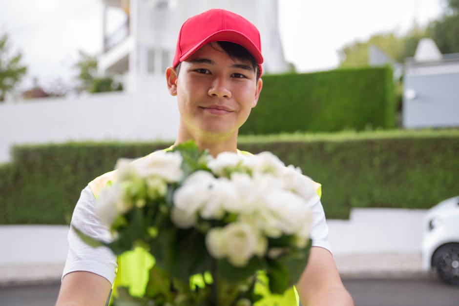 A smiling delivery person wearing a red cap and a high-visibility vest, holding a bouquet of fresh white flowers with green leaves, in an outdoor residential setting with greenery, white buildings, an
