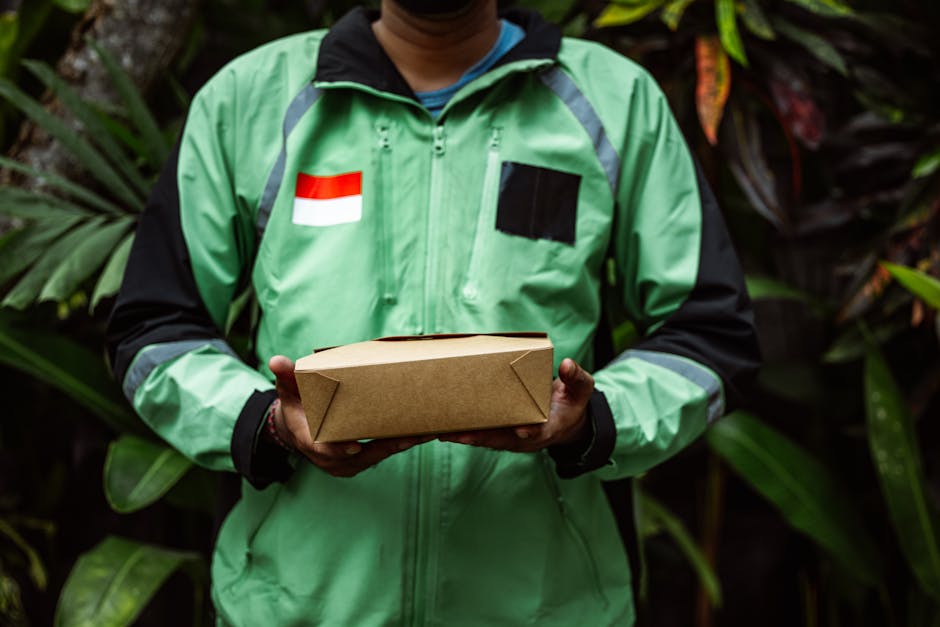 A delivery person wearing a green jacket with black accents, an Indonesian flag patch, and a black patch on the chest, is holding a rectangular cardboard parcel with both hands. The individual is stan