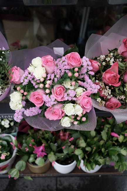 A floral arrangement featuring a bouquet of fresh pink roses, white carnations, and small pink and white filler flowers, wrapped in light purple translucent paper. The flowers are arranged in a rounde